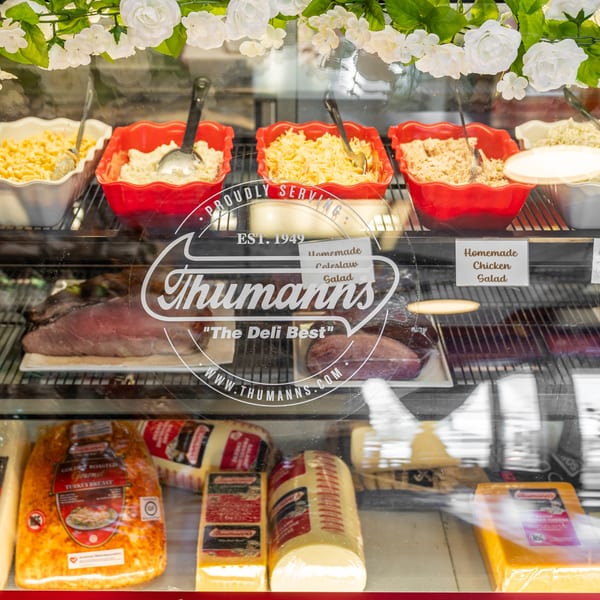 Deli display case with salads, meats, and cheeses, featuring the Thumann's logo.