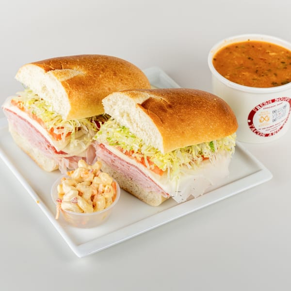 Two deli sandwiches with lettuce, tomato, and meat, served with macaroni salad and a cup of soup.