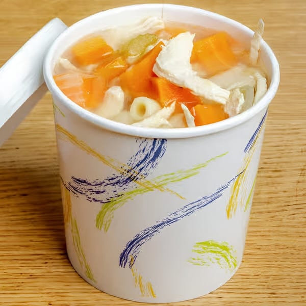 Chicken noodle soup with carrots and celery in a colorful to-go cup on a wooden table.