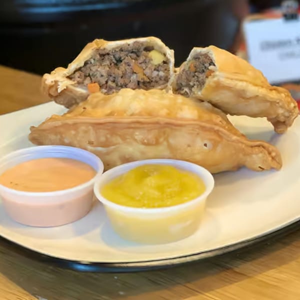 Three golden-brown empanadas, one cut open to show a savory meat and vegetable filling, with two dipping sauces.