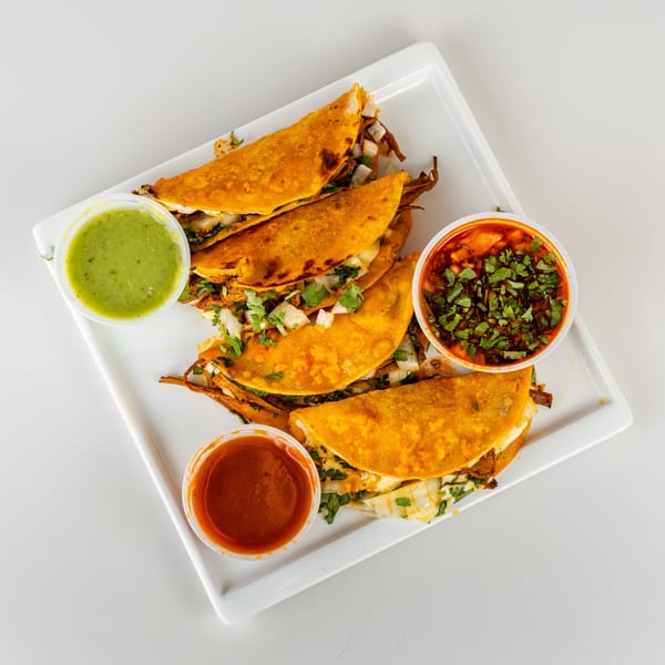 Four golden-brown birria tacos with cilantro and onions, served with three dipping sauces on a white plate.
