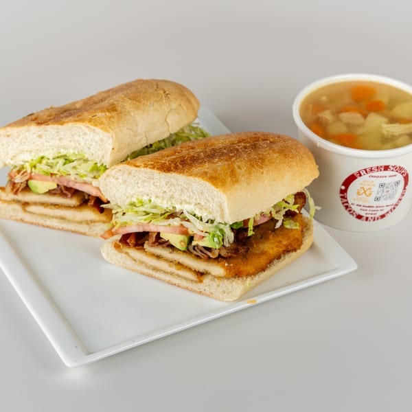 Two halves of a crispy chicken sandwich with lettuce, tomato, and avocado, next to a cup of chicken noodle soup.